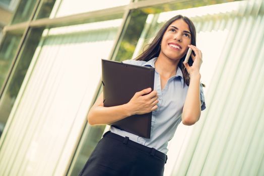 Young beautiful business woman with planner in her hands, standing in front of office building and talking on mobile phone