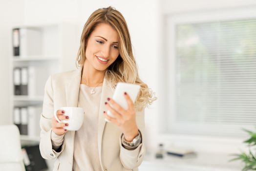 Beautiful young businesswoman relaxing in the office with coffee and using phone.