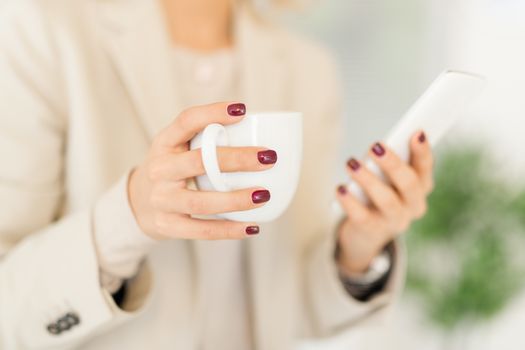 Close-up of female hand with painted nails holding coffee and typing on smart phone.