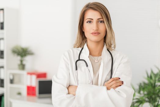 Young cute female doctor with stethoscope standing in her consulting room and looking at the camera.