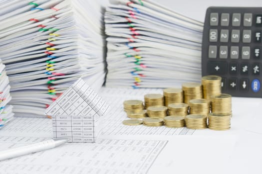 House and silver pen on finance account have blur step pile of gold coins with calculator and pile of paperwork as background.