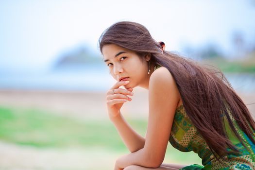 Beautiful biracial Asian Caucasian teen girl sitting at tropical beach, thinking, with ocean in background