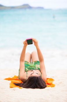 Beautiful biracial Asian Caucasian teen girl lying on orange blanket at beach, arms overhead looking at cellphone. Ocean in background