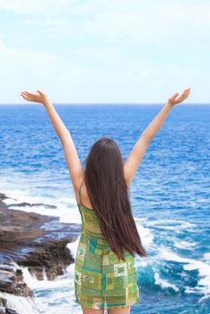 Beautiful biracial Asian Caucasian teen girl arms raised upwards in praise by blue ocean waters along rocky shore