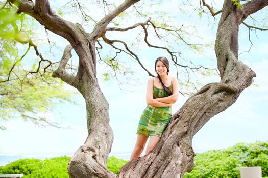 Beautiful biracial Asian Caucasian teen girl standing on branches of large branching tree in Hawaii with ocean in background 