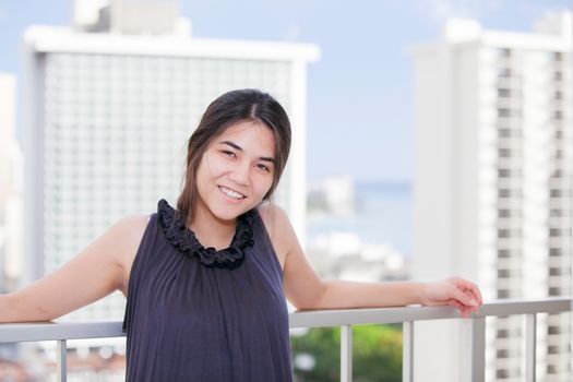 Beautiful biracial Asian Caucasian teen girl standing on outdoor high rise patio deck, leaning on railing with urban scene of high buildings and ocean landscape in background