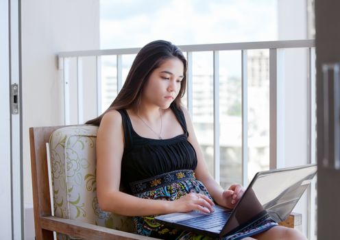 Beautiful biracial Asian Caucasian teen girl sitting on outdoor high rise patio studying or working on a laptop computer with urban city background