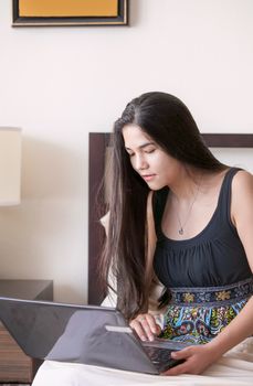 Beautiful biracial Asian Caucasian teen girl sitting in bed studying or working on a laptop computer in bedroom