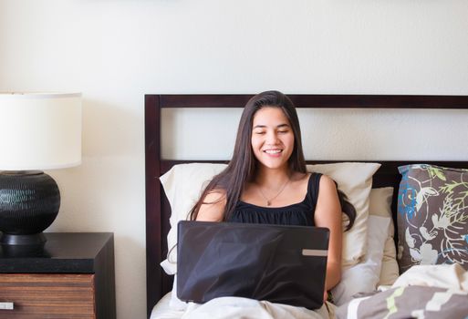 Beautiful biracial Asian Caucasian teen girl sitting in bed studying or working on a laptop computer in bedroom