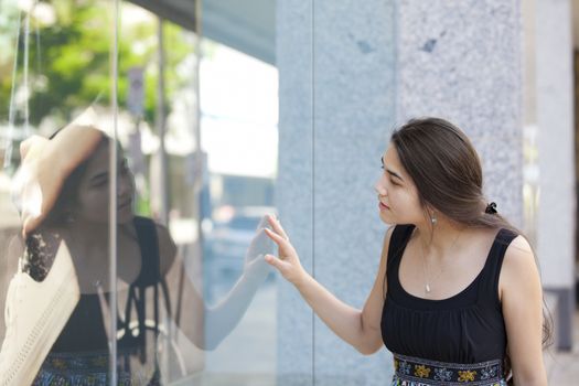 Beautiful biracial Eurasian Asian Caucasian teen girl standing in front of department store window looking in, window shopping