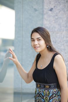 Beautiful biracial Eurasian Asian Caucasian teen girl standing in front of department store window looking in, window shopping