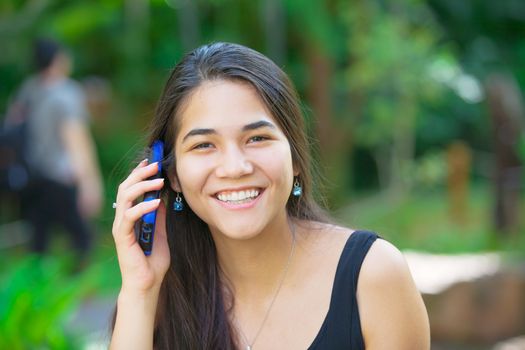 Beautiful biracial Asian Caucasian teen girl talking on cell phone outdoors with green foliage in background
