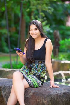 Beautiful biracial Asian Caucasian teen girl sitting on rock in tropical setting looking at cellphone, palm trees in background