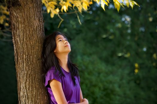 Biracial Asian  Caucasian teen girl leaning against tree with autumn leaves looking up, thinking