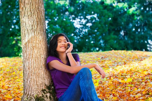 Biracial  teen girl sitting with back to tree using cell phone, autumn leaves on ground