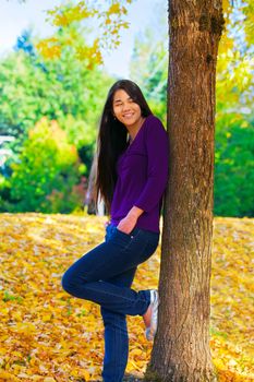 Beautiful biracial Asian Caucasian teen girl leaning against tree with autumn leaves on ground in background