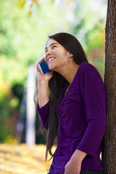 Beautiful biracial Asian Caucasian teen girl leaning against tree talking happily on cell phone, autumn leaves on ground