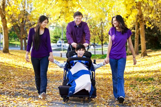 Multiracial family with disabled child in wheelchair walking among autumn leaves 