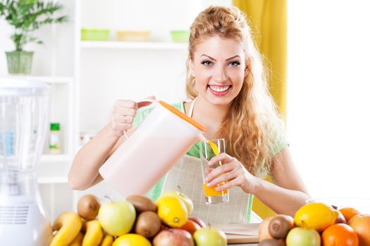 Beautiful young woman pouring orange juice at glass in a kitchen. Looking at camera.