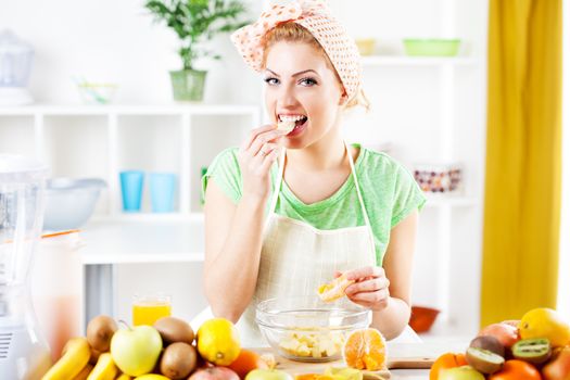 Beautiful young woman preparing and eat orange for fruit salad in a kitchen. Looking at camera.