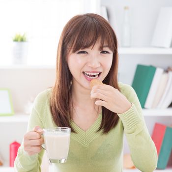 Portrait of happy Asian girl eating cookies and having soymilk as breakfast. Young woman indoors living lifestyle at home.