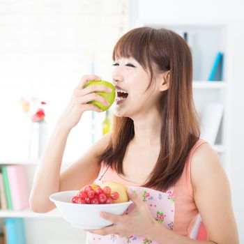 Happy Asian housewife with apron eating green apple, holding bowl of fresh fruits. Young woman indoors living lifestyle at home.