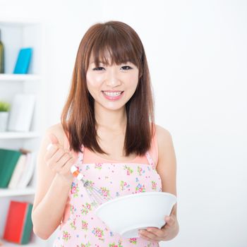 Happy Asian housewife with apron preparing food, using manual egg mixer and bowl. Young woman indoors living lifestyle at home.