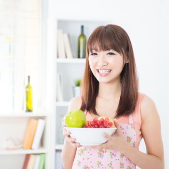 Happy Asian housewife with apron holding a bowl of fresh fruits. Young woman indoors living lifestyle at home.