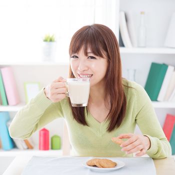 Portrait of happy Asian girl drinking soymilk and having cookies as breakfast. Young woman indoors living lifestyle at home.