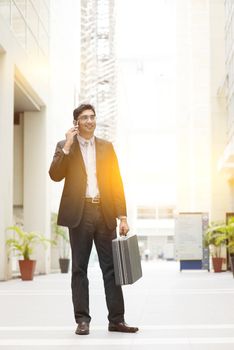 Portrait of fullbody Asian Indian business man using smartphone, standing outside modern office building block, beautiful golden sunlight at background.