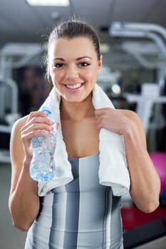 Cute Young girl standing and resting in modern gym and holding a bottle of water and towel. Looking At Camera.