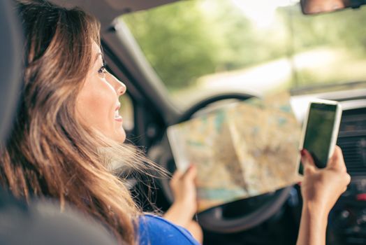 Young smiling beautiful woman sitting in car, holding map and smart phone. Rear view.