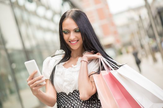 Happy girl in shopping walking out of shopping mall with bags and looking at smart phone.