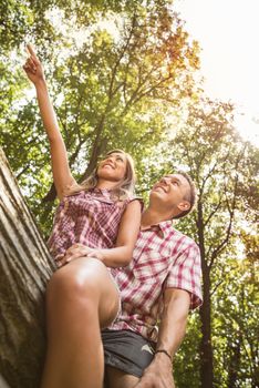 Beautiful young couple sitting on fallen tree in the forest and enjoying. Girlfriend pointing with finger far away.