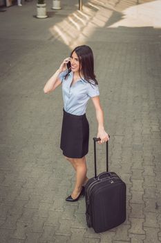 Young beautiful business woman standing outdoor with suitcase ready for a business trip and talking on mobile phone.