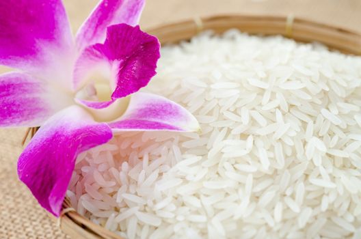 Raw rice with violet orchid flower in weave basket on sack background.