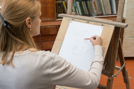 A young girl draws a pencil on the easel with a portrait sculpture in the artists studio