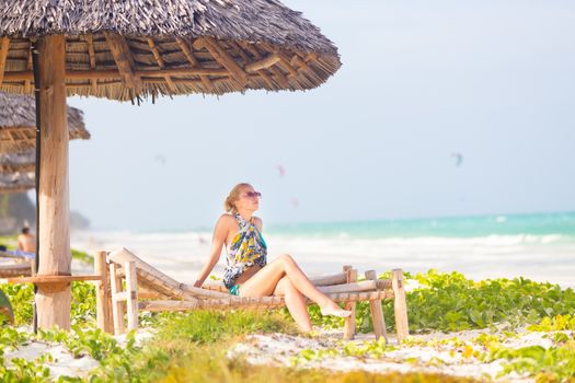 Women sunbathing on dack chair under wooden umbrella on stunning tropical beach. Kiteboarders turquoise blue lagoon of Paje beach, Zanzibar, Tanzania in the background.