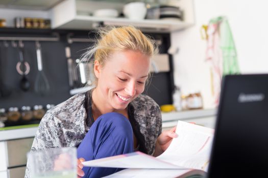 Female student in her casual home clothing working and studying remotly from her small flat in the morning. Home kitchen in the background. Great flexibility of web-based courses and study programmes.