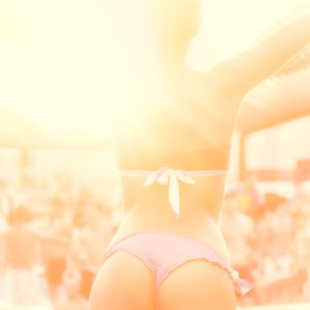 Sexy hot girl wearing brazilian bikini dancing on a beach party event in sunset. Crowd dancing and partying at poolside in background. Summer electronic music festival. Hot summer party vibe.