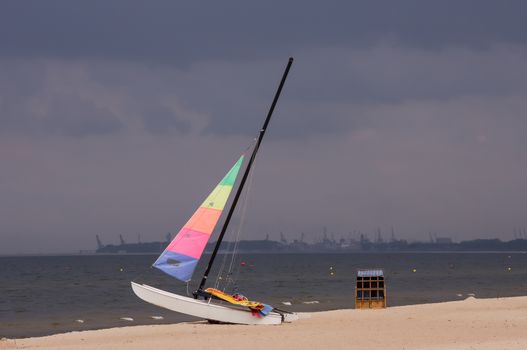 Colorful sailboat on the beach in Sopot, Poland, with the stormy clouds and Gdansk Shipyard in the background.