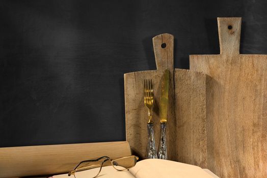 Empty blackboard on the wall with cutting boards, silver cutlery, recipe book, glasses and rolling pin. Concept of rustic cuisine