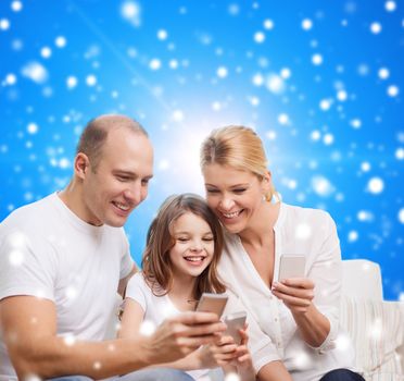 family, christmas holidays, technology and people concept - smiling mother, father and little girl with smartphones over blue snowy background