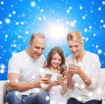 family, christmas holidays, technology and people concept - smiling mother, father and little girl with smartphones over blue snowy background