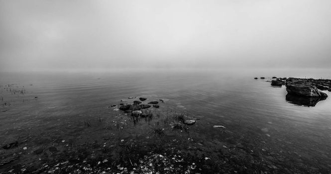 Thick dense fog enveloping Ottawa River shoreline - bright diffusion in low misty cloud, rocks in still water on mid-morning sunrise.