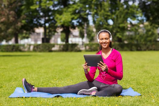 fitness, park, technology and sport concept - smiling african american woman with tablet pc computer and headphones on mat outdoors