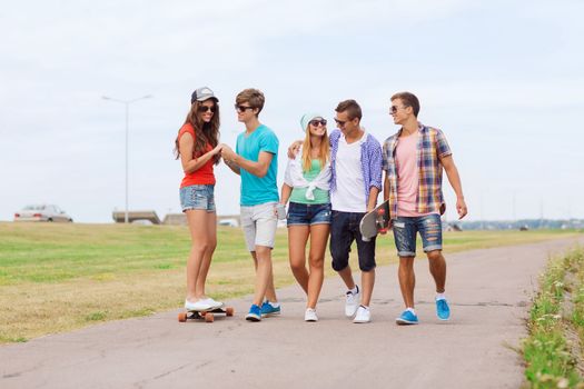 holidays, vacation, love and friendship concept - group of smiling teenagers walking and riding on skateboards outdoors