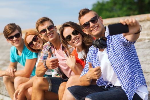 friendship, summer, technology, gesture and people concept - group of smiling friends with smartphone and headphones making selfie outdoors