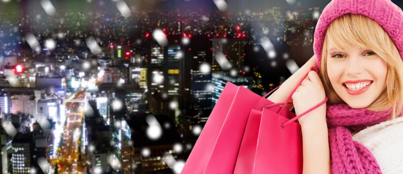 happiness, winter holidays, christmas and people concept - smiling young woman in hat and scarf with pink shopping bags ove snowy night city background