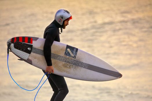 CAPE SOLANDER, AUSTRALIA - AUGUST 27, 2015; Surfer wearing a gath surf helmet and wetsuit and carrying a surfboard at sunrise while  at the notorious reef break at Cape Solander where only the best and bravest surf.  The helmet has many scratches